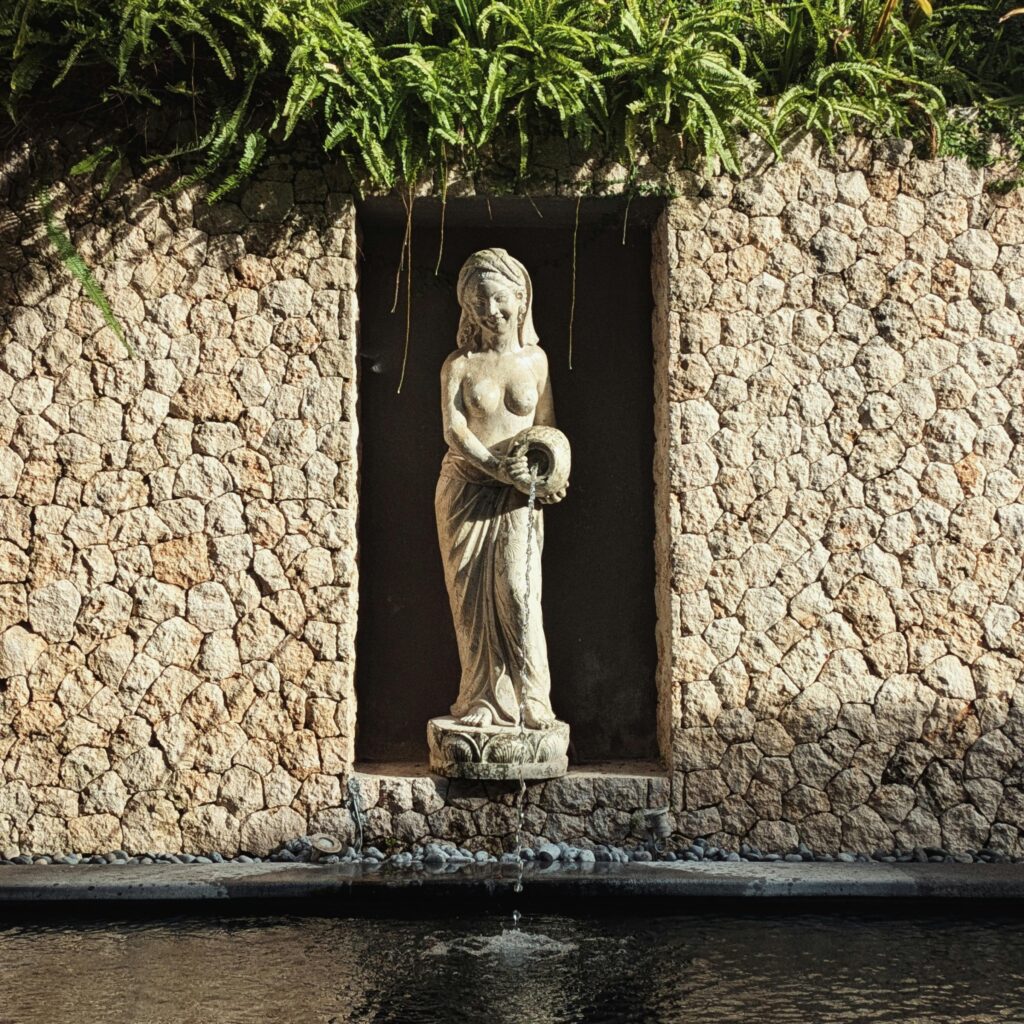 Stone statue of a woman in a garden alcove, surrounded by lush ferns and water feature.
