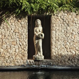 Stone statue of a woman in a garden alcove, surrounded by lush ferns and water feature.