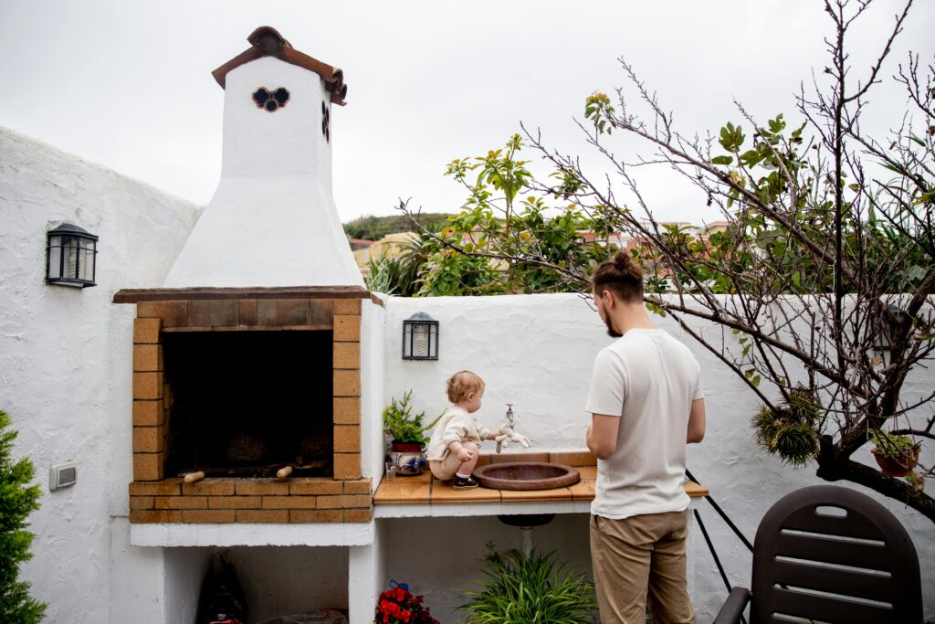 Man wearing casual clothes exploring outdoor kitchen with fireplace and sink together with cute toddler kid in backyard near white stone fence during daytime at countryside