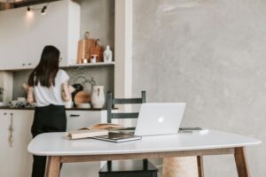 Laptop on a white table in a stylish home office, with a person in the kitchen.
