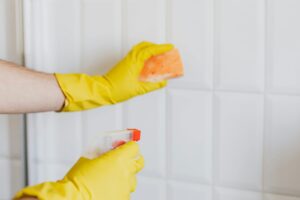 A person wearing yellow gloves cleans bathroom tiles with a sponge and spray bottle.