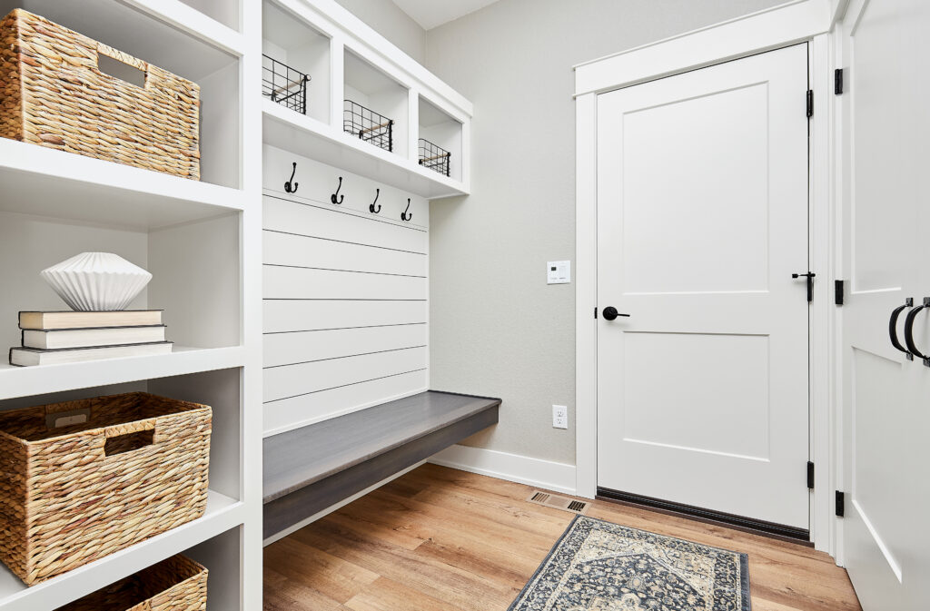 A clean, organized garage entryway mudroom with built-in storage cabinets, a wooden bench, hooks hanging coats, and durable wood flooring.