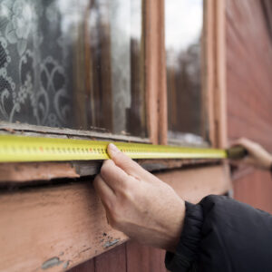 A person using a yellow tape measure to find the dimensions of a weathered, peeling wooden window frame on an older home.