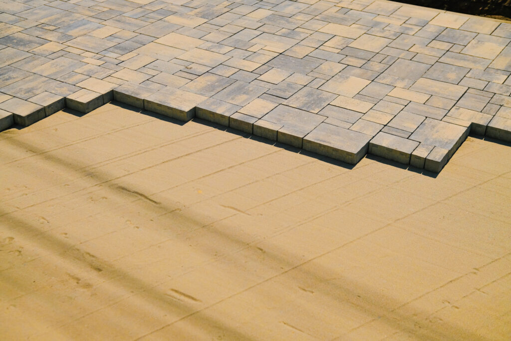 A close-up, high-angle shot of grey and tan multi-sized pavers being installed on a perfectly smooth, leveled sand base.
