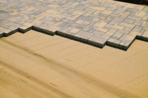 A close-up, high-angle shot of grey and tan multi-sized pavers being installed on a perfectly smooth, leveled sand base.