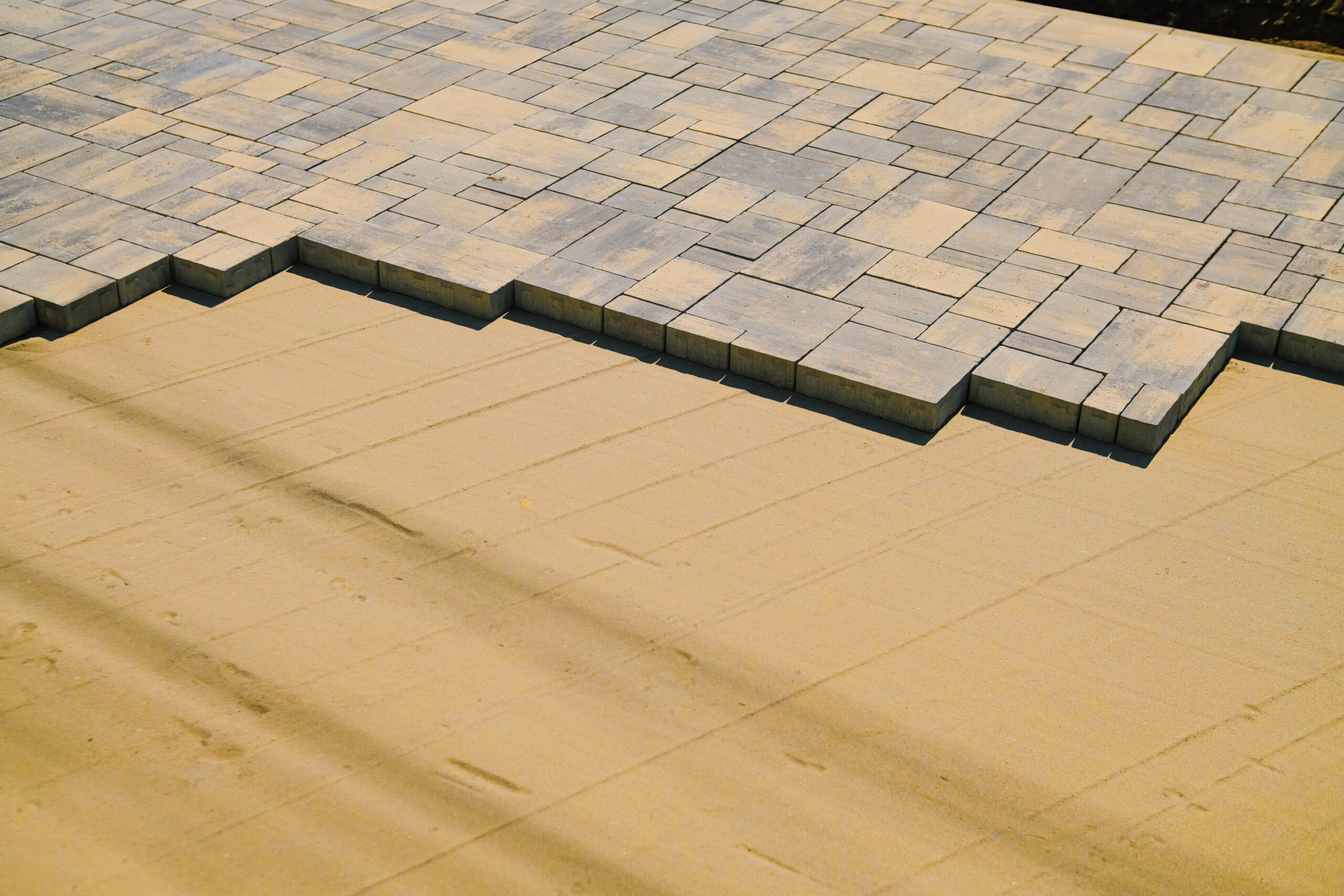 A close-up, high-angle shot of grey and tan multi-sized pavers being installed on a perfectly smooth, leveled sand base.