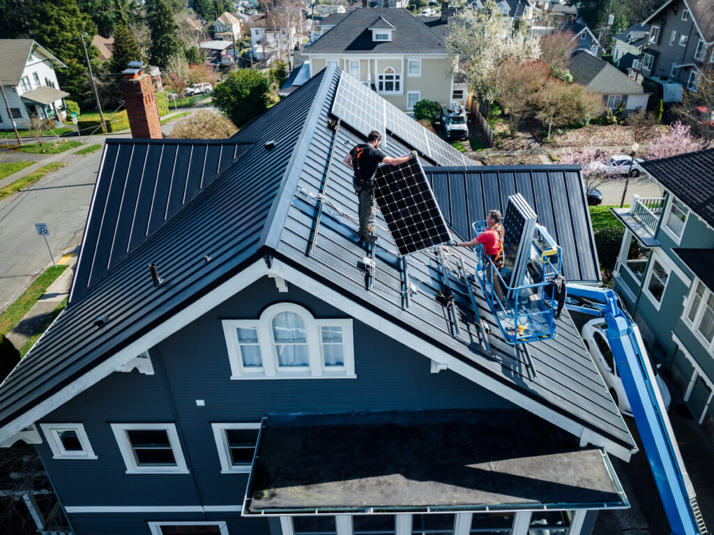 An aerial drone view of two contractors installing solar panels onto a dark grey standing-seam metal roof of a multi-story blue house using a cherry picker lift.