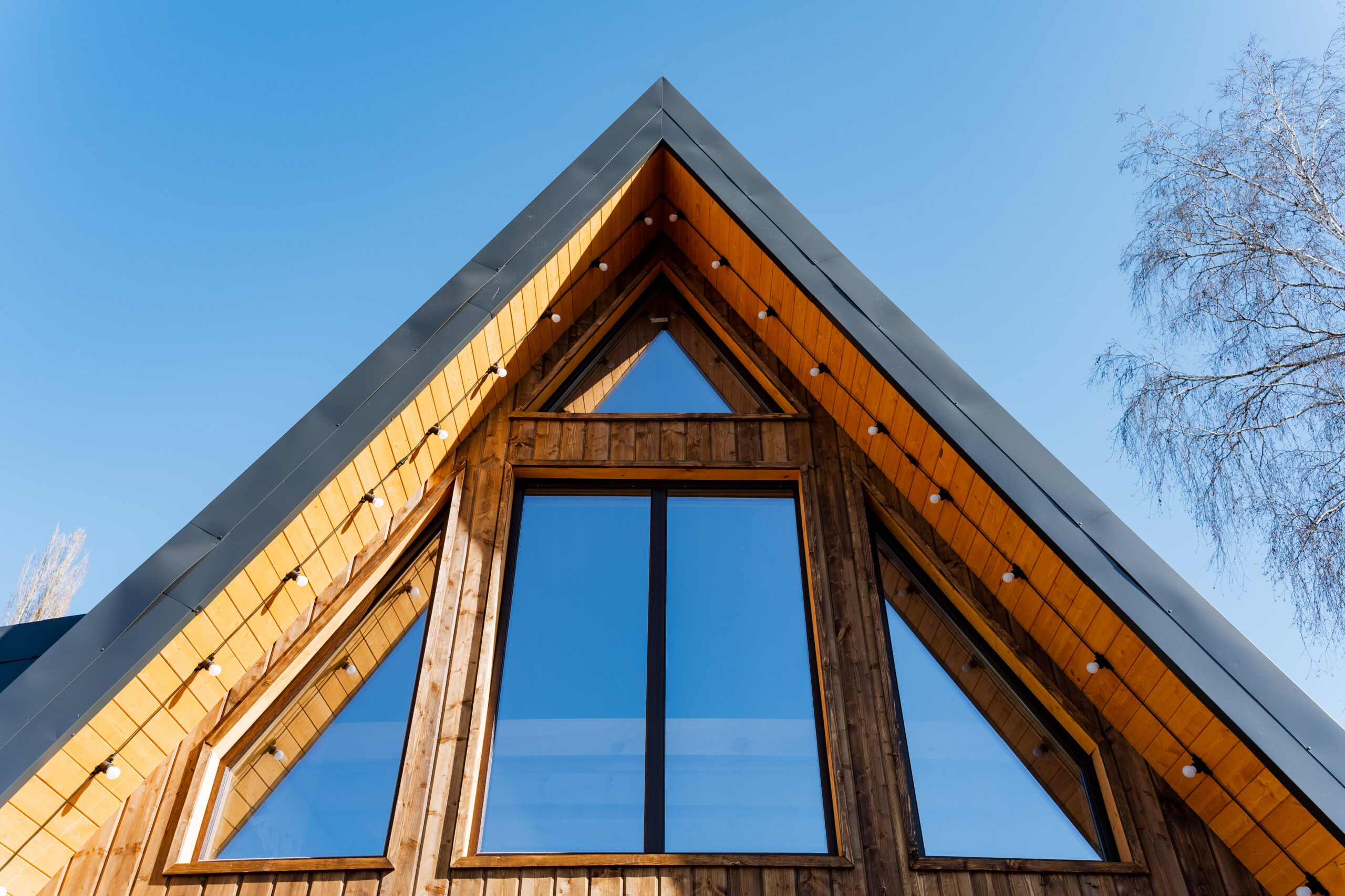 A low-angle, symmetrical view of a modern A-frame wooden cabin against a clear blue sky. The facade features large triangular and rectangular windows that reflect the sky, framed by light-toned wood siding and a dark metal roofline with integrated string lighting.