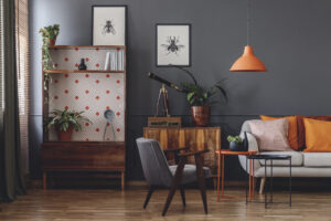 A modern living room with a dark charcoal wall featuring a blend of mid-century modern and eclectic vintage decor. The room includes a tall wooden hutch with a geometric patterned backing, a grey velvet armchair, and a white sofa accented with orange pillows. An orange pendant light hangs above a nesting side table, while botanical sketches of insects and a vintage brass telescope on a sideboard add a curated, scholarly feel.