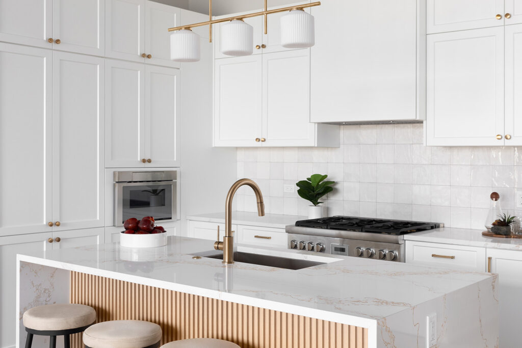 White kitchen with a quartz waterfall island, brass faucet, fluted wood island panel and pendant lighting