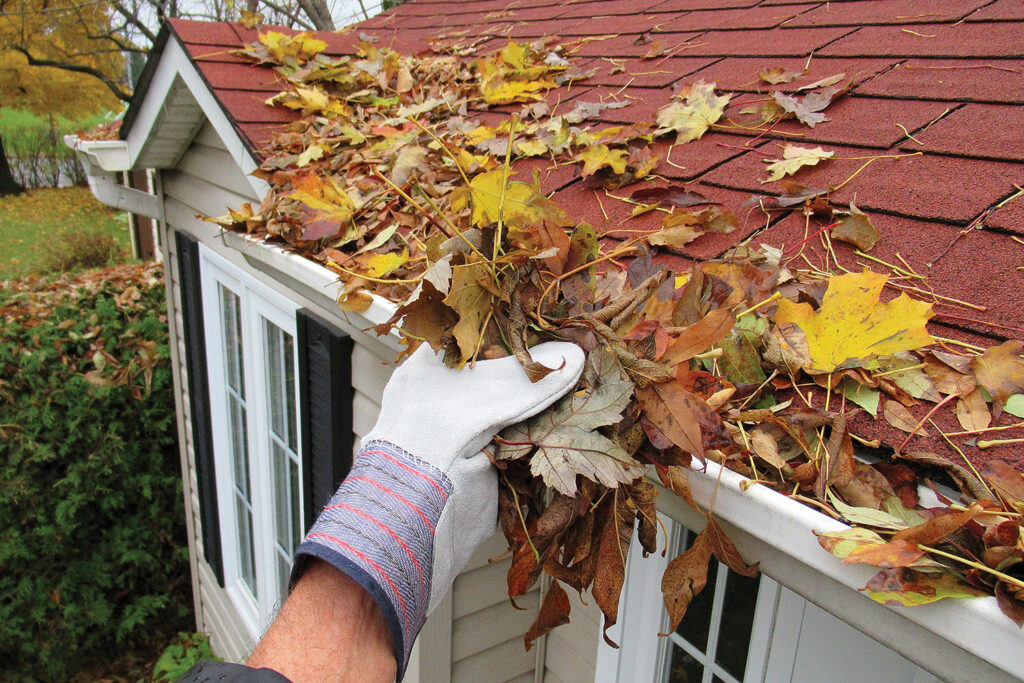 Gloved hand clearing wet leaves from a home gutter along a shingled roofline in fall