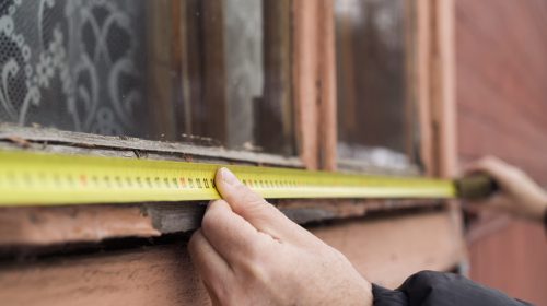 A person using a yellow tape measure to find the dimensions of a weathered, peeling wooden window frame on an older home.