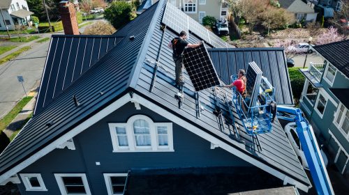 An aerial drone view of two contractors installing solar panels onto a dark grey standing-seam metal roof of a multi-story blue house using a cherry picker lift.