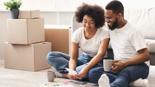 Renovation. Black Couple Planning To Paint Walls Choosing Color Together With Palettes Sitting On Floor Among Moving Boxes In New House. Family, Real Estate And Apartment Improvement Concept