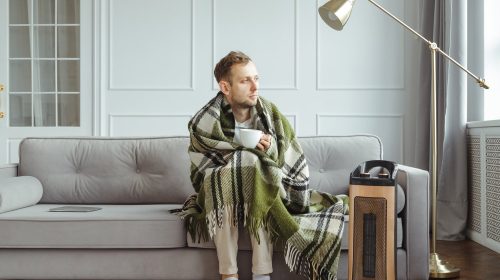 Young man in wrapped in plaid in warm clothes with cup of tea at home. Concept of heating season