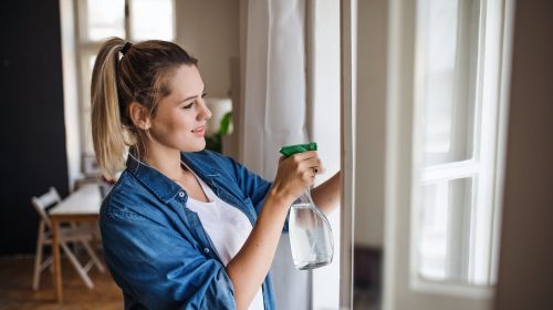 A young woman standing indoors at home, cleaning windows.