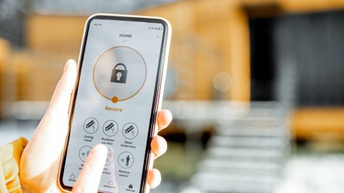 Holding phone with launched alarm app on the house background outdoors. Concept of controlling and managing home security from a mobile device