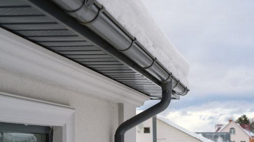 Corner of house with new roof made of gray metal tiles and gutter covered with thick layer of snow in winter. Metal Downpipe system, Guttering System, External downpipes and drainage pipes
