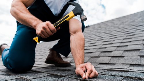 cropped view of handyman in uniform holding hammer while repairing roof