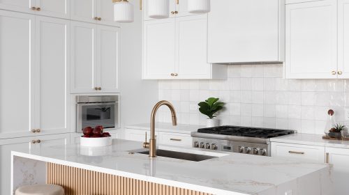 White kitchen with a quartz waterfall island, brass faucet, fluted wood island panel and pendant lighting