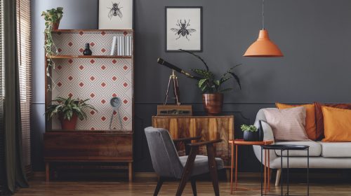 A modern living room with a dark charcoal wall featuring a blend of mid-century modern and eclectic vintage decor. The room includes a tall wooden hutch with a geometric patterned backing, a grey velvet armchair, and a white sofa accented with orange pillows. An orange pendant light hangs above a nesting side table, while botanical sketches of insects and a vintage brass telescope on a sideboard add a curated, scholarly feel.