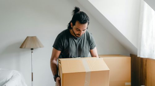 A man with a ponytail lifts a cardboard box in an attic room, suggesting relocation.