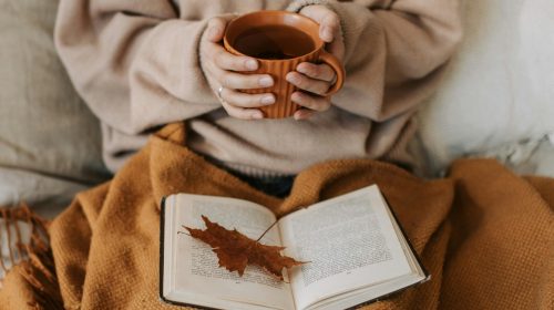 A woman enjoys a cozy autumn day, holding a tea mug over an open book with a maple leaf.